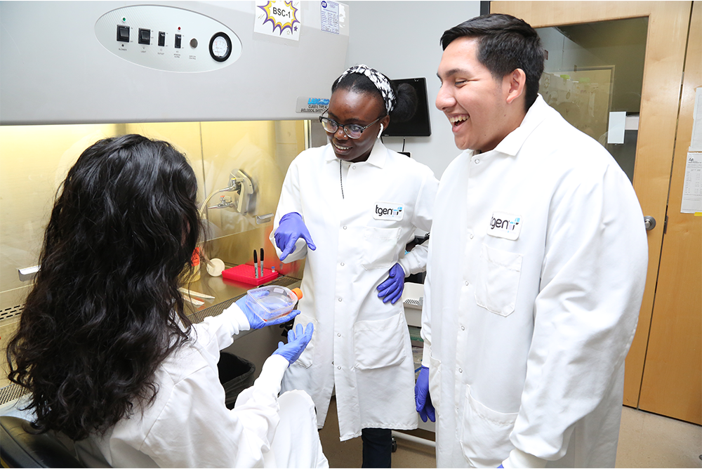 Three TGen students wear white lab coats in an enclosed lab